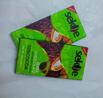 Two green-wrapped bars of Sekoe Chocolates, labeled as "Dark Chocolate 70% Cocoa," placed on a white textured surface. The packaging features images of cocoa beans and open cocoa pods.