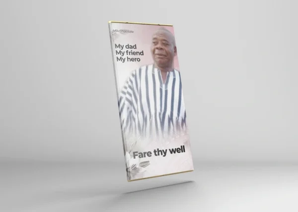A rectangular custom-wrapped chocolate bar designed as a funeral tribute. The wrapper features a photo of an elderly man wearing a traditional striped outfit. The text reads: “My dad, My friend, My hero” at the top and “Fare thy well” at the bottom, accompanied by soft floral accents. The background is light with a gentle pink hue, giving the design a respectful and heartfelt tone