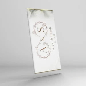 A custom wedding chocolate bar with an elegant white wrapper. The design features floral wreaths around the initials "A & S" and includes the names "Alex & Sarah" along with the wedding date "05.24.24" written vertically in a delicate serif font. Soft floral imagery decorates the top edge, giving the design a minimalist and romantic feel.